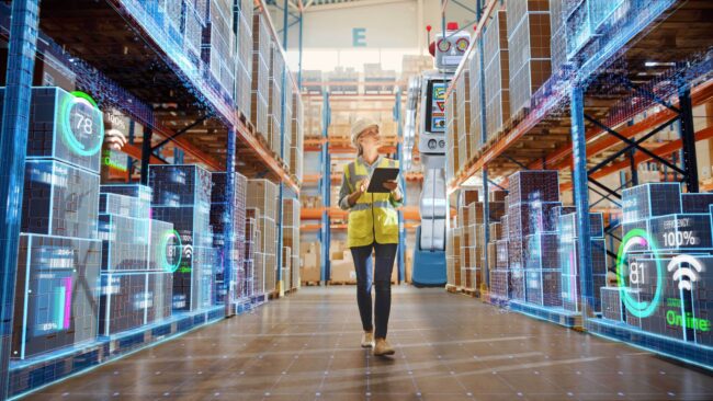 Warehouse worker with neon yellow vest walking between aisles of boxes. She is being watched by a robot.