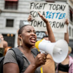 Black woman shouting into a megaphone in front of protesters holding a sign that says "Fight today for a better tomorrow"