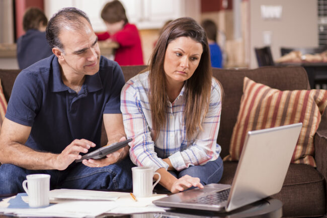 Latino man and woman struggle to pay their monthly bills. They are calculating expenses versus budget income and are upset. Many invoices, laptop on living room coffee table. Children in kitchen background. Frustration among middle-class people. Imagery for home finances, recession
