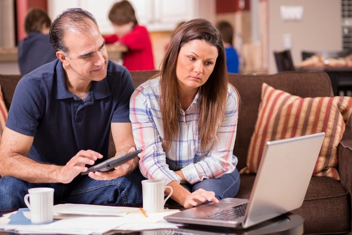 Latino man and woman struggle to pay their monthly bills. They are calculating expenses versus budget income and are upset. Many invoices, laptop on living room coffee table. Children in kitchen background. Frustration among middle-class people. Imagery for home finances, recession