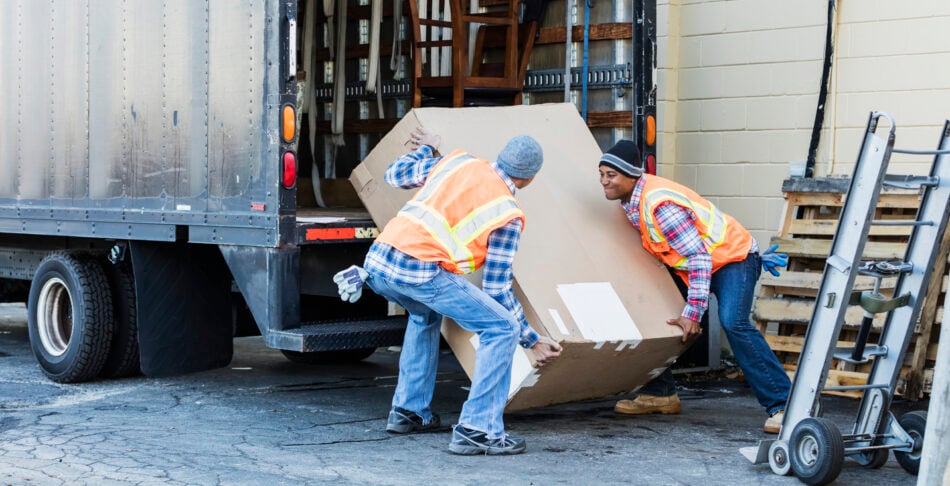 Two multi-ethnic mature workers in their 40s at the back of a truck, loading or unloading a large cardboard box. The men are wearing plaid shirts, reflective vests and jeans.