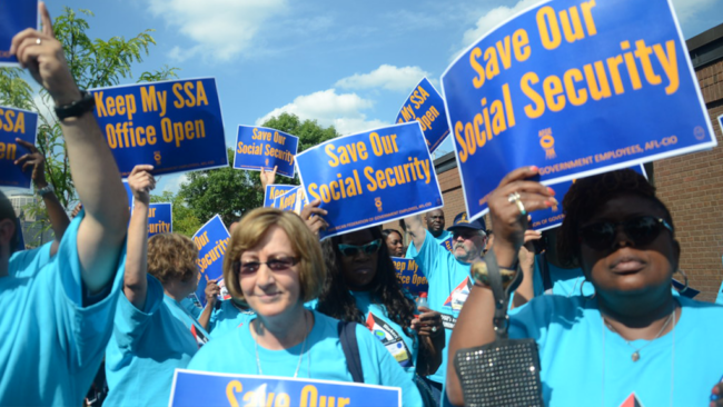 Women in bright teal t-shirts marching on a blue-sky day, holding up blue posters that have yellow text reading "Save our social security"