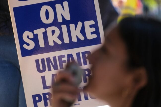 picture of people at a protest holding a white sign with blue writing that says "On Strike unfair labor practices"